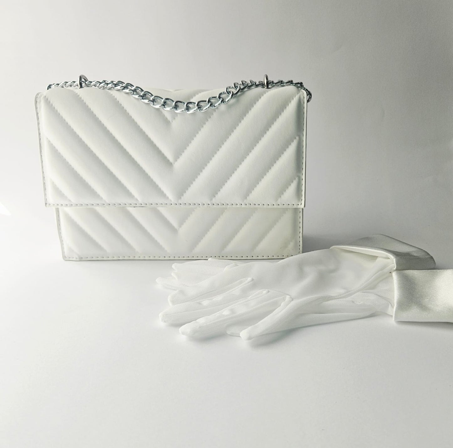 White handbag with a chain strap and white gloves on a light gray background